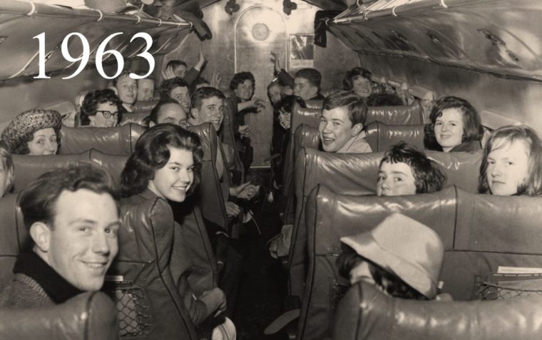 A group of children sitting on an aeroplane look backwards over their chairs at the camera.