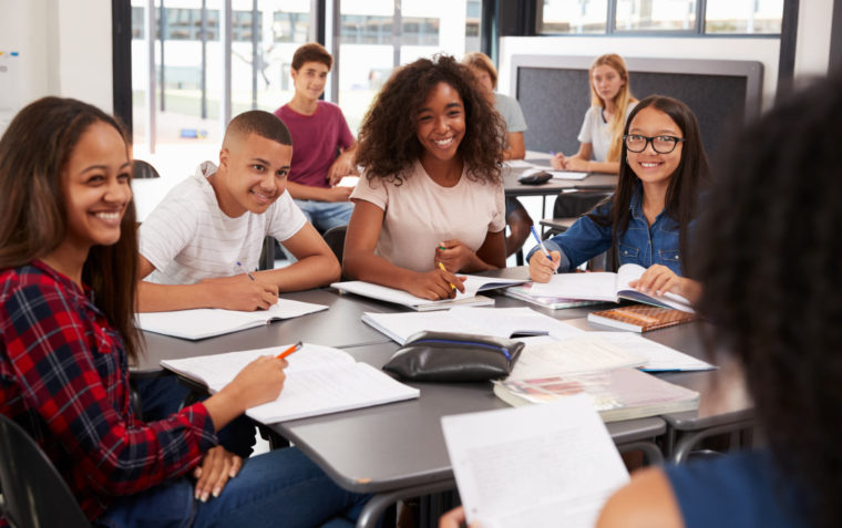 A group of secondary school students sitting inside a classroom and smiling to their teacher.