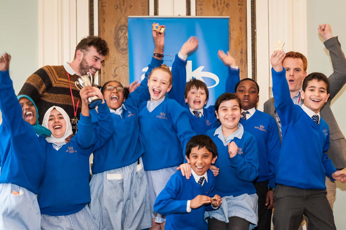 A group of cheering primary school pupils with two teachers at the back. Two of the girls are wearing headscarves and one is holding a trophy in her hand.