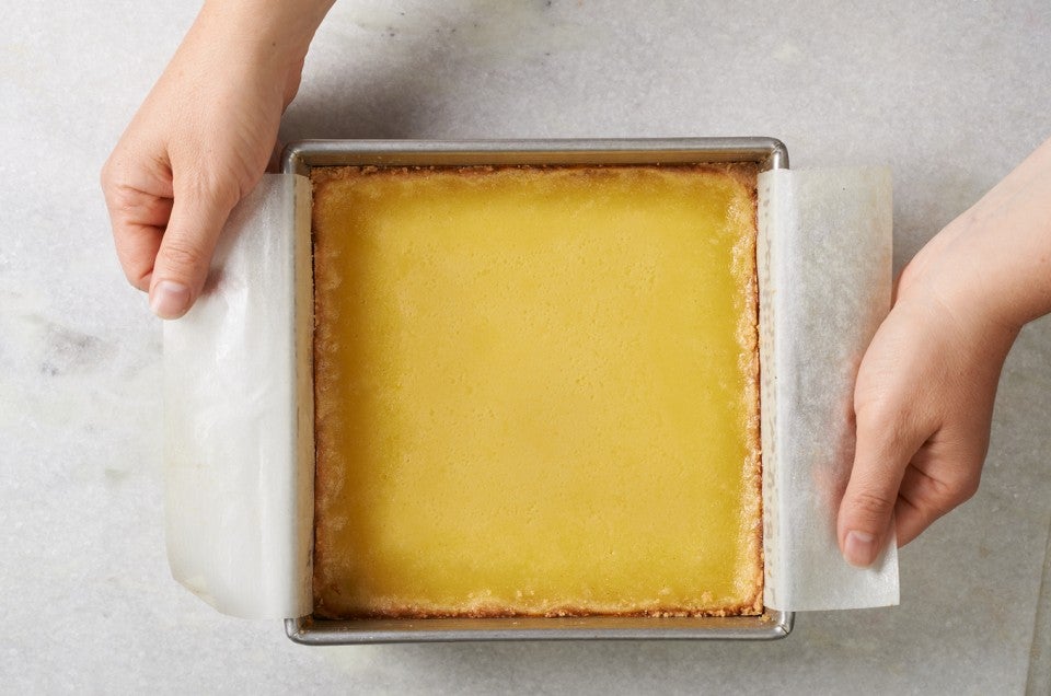 Hands using parchment paper sling to remove lemon bars from square pan