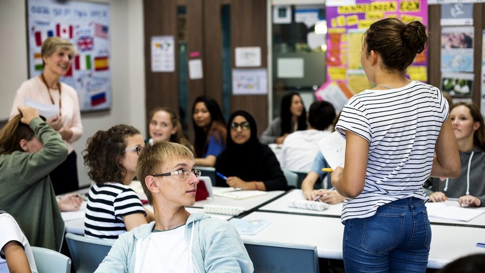 A group of students sitting in a classroom. A young female students is standing in the back of the room while speaking directly to the female teacher also standing in front of the room with handouts in her hand.