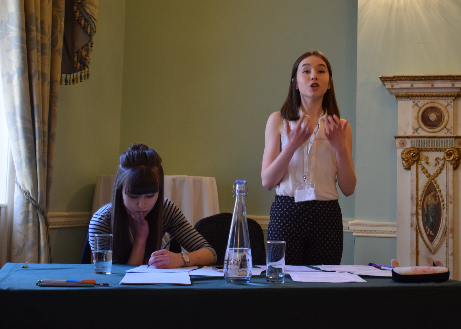 A young pupil passionately delivers her speech while her team mate is writing down her notes