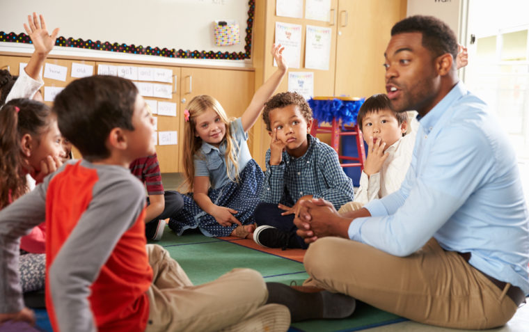 A primary school male teacher and a group pupils (boys and girls) sitting in circle on the floor. A boy and a girl raised their hands in the air.