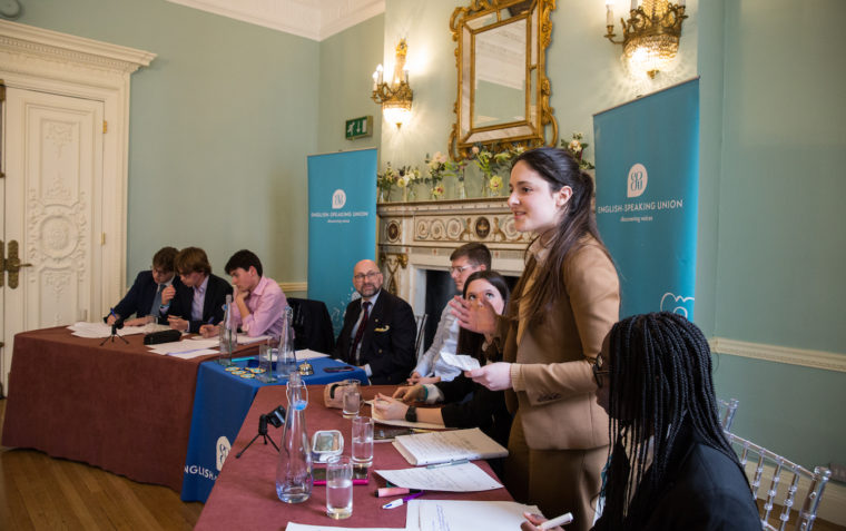 A pupil standing and delivering her speech at an ESU debating and public speaking competition while her teammates watch her and her opponents listen and write down their notes