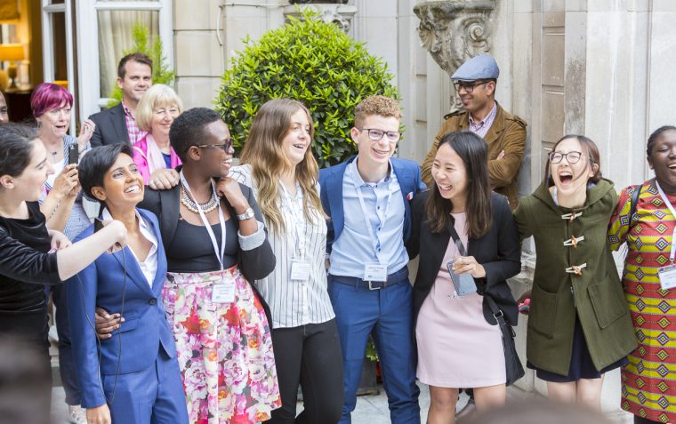 A group of secondary school students (male and female) standing outside a building smiling and laughing with hands across each other.