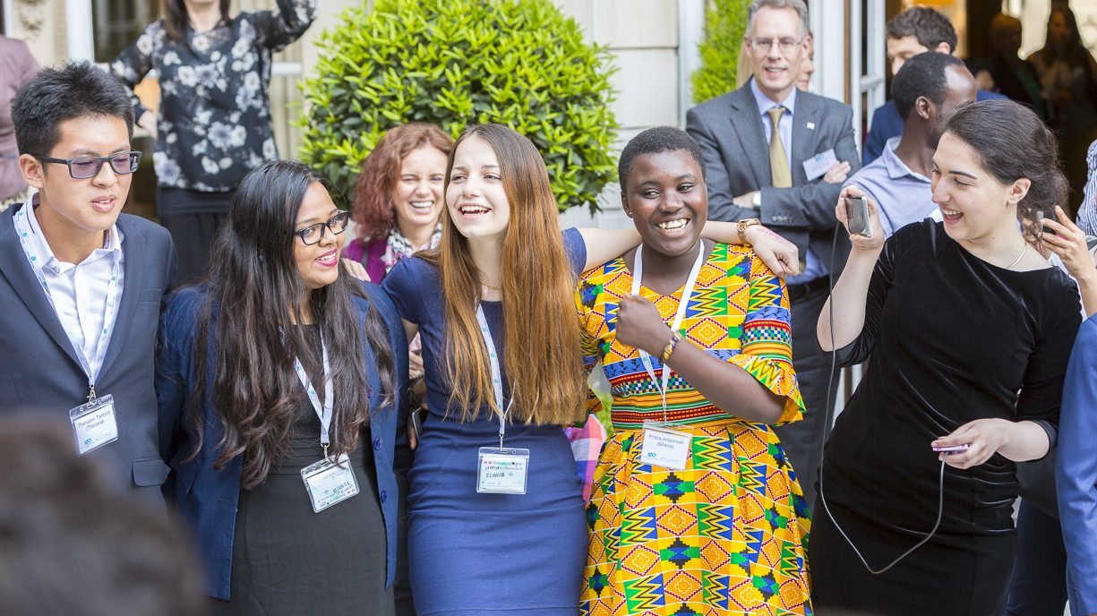 A group of international secondary school students dressed in different attires standing closed together outside a building and smiling.