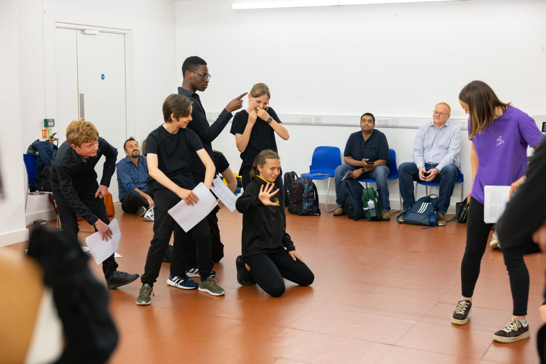 A group of secondary school students rehearsing one of Shakespeare's play in the centre stage with observers sitting at the back. 