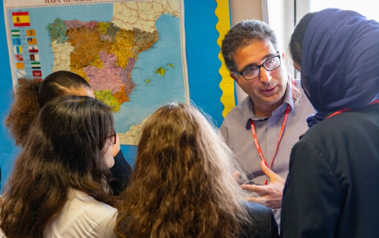 Four female secondary school students listening to the male teacher speaks. The teacher is wearing lens glasses and one of the female student is wearing a headscarf. A map of the world is hanging on the wall in the background.
