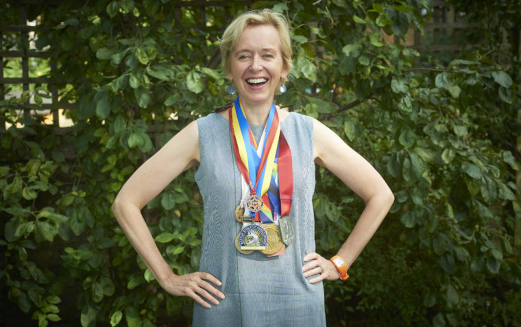 A picture of Rosie Millard OBE and former chair of Hull city of culture wearing her medals, smiling with arms akimbo and standing in front of a little bush.