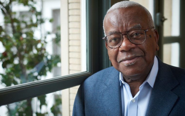 A close-up photograph of Sir Trevor McDonald next to a window with trees behind