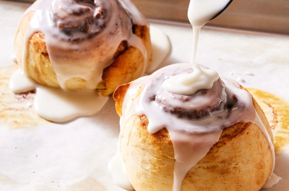 A small batch of two cinnamon rolls on a quarter sheet pan being frosted with icing - select to zoom