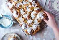 A batch of soft cinnamon rolls on a cooling rack with a hand reaching for a roll